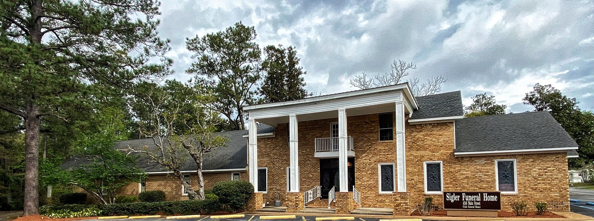 Brick house with white columns and a porch under cloudy sky, surrounded by trees