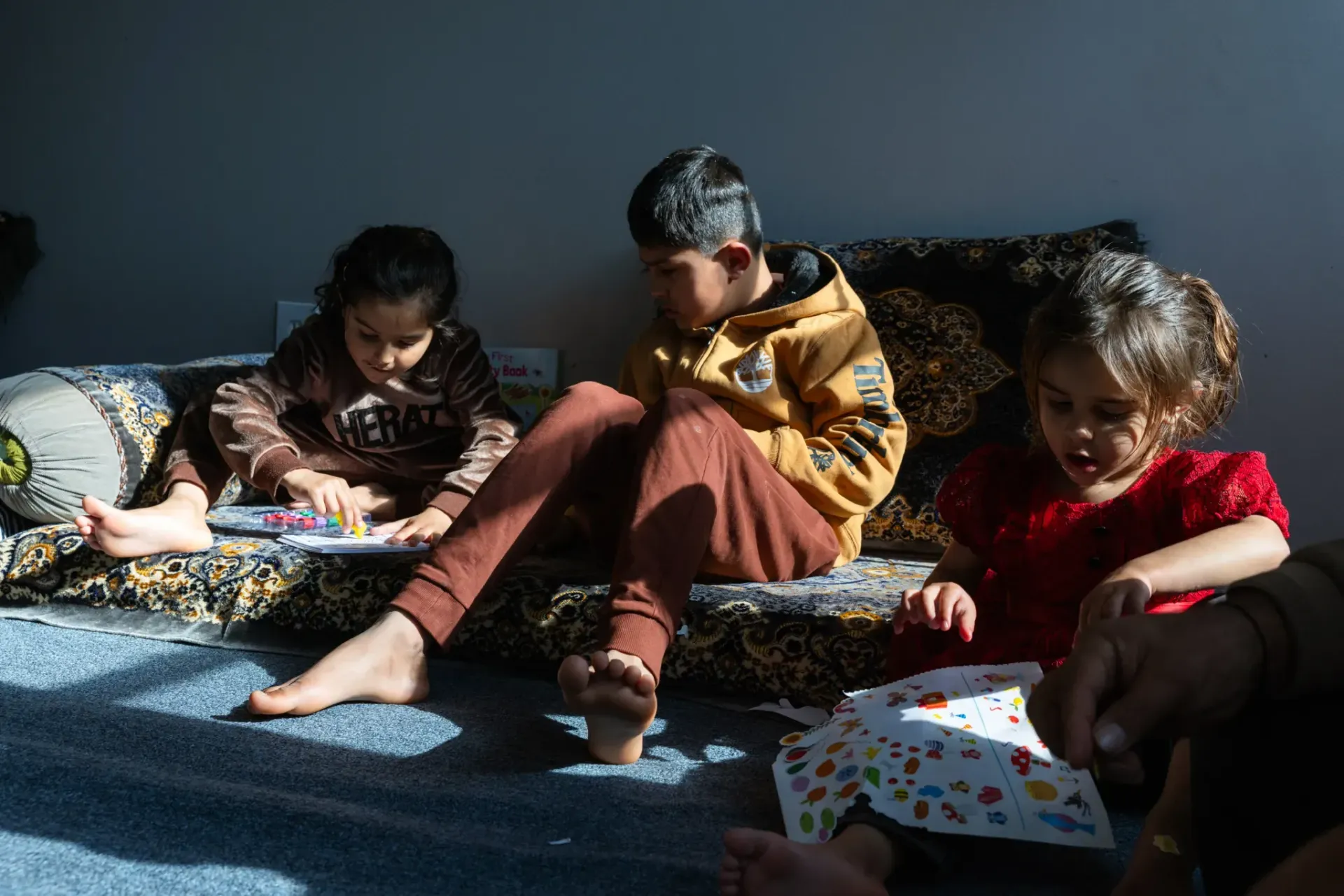 Three children sit on floor cushions in a sunlit room, focusing on books and papers in their laps.