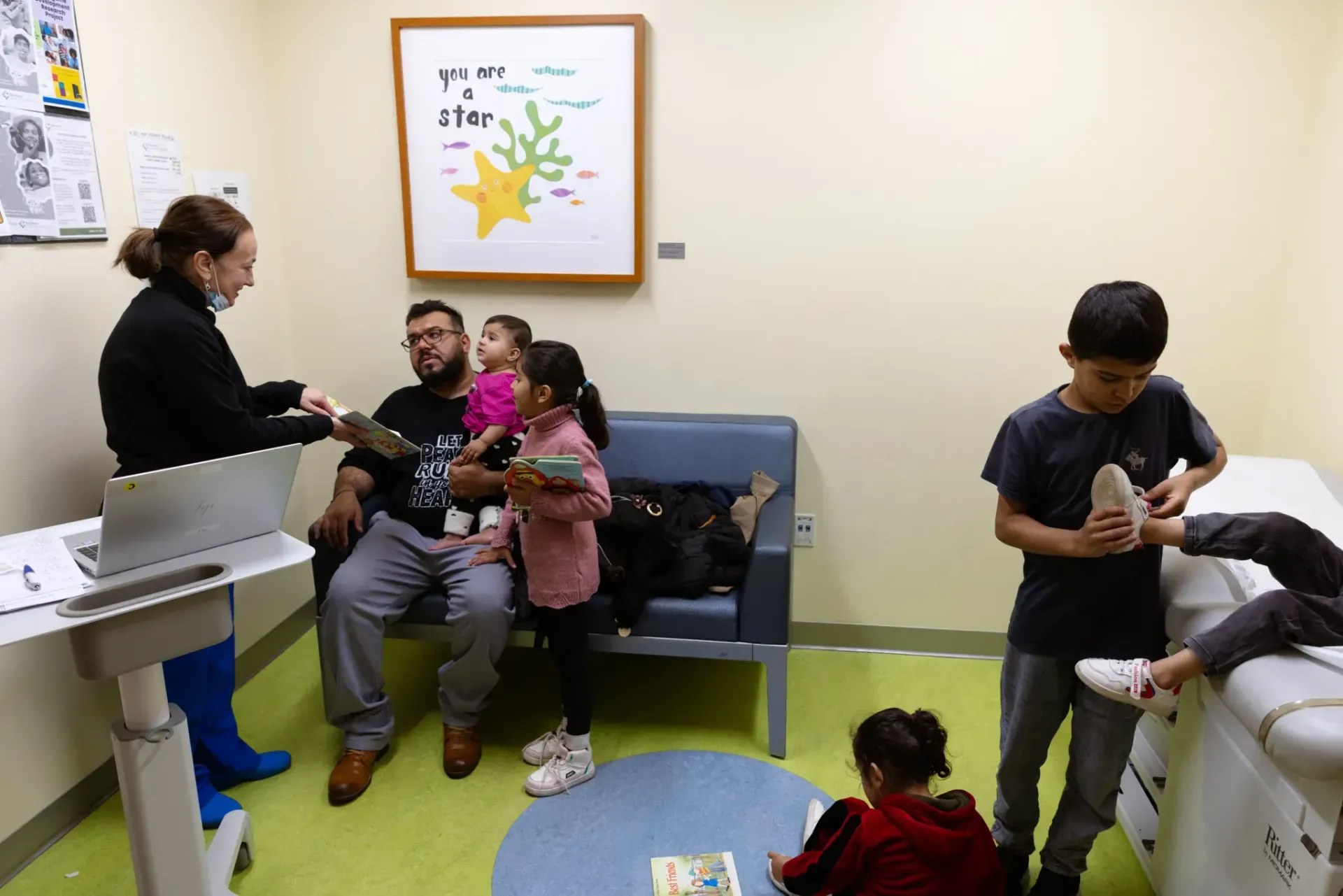 A medical professional interacts with a family in a clinical examination room with light green flooring and a wall poster.