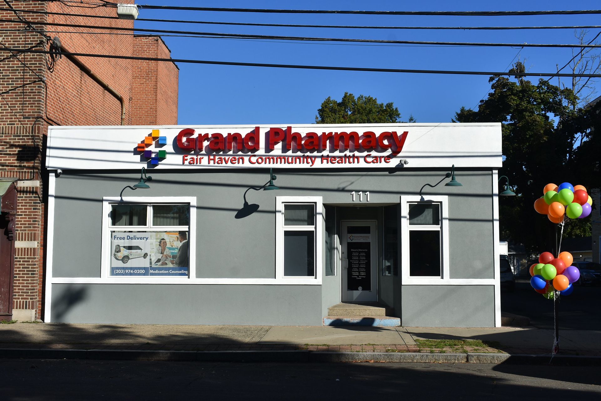 Grand Pharmacy storefront with gray facade, red sign, and colorful balloons outside