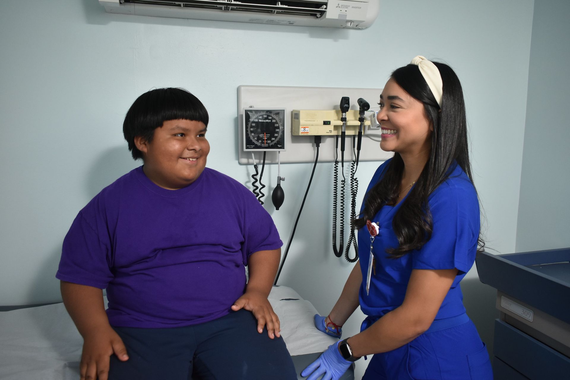 Nurse in blue smiles while checking a seated patient in a medical exam room.