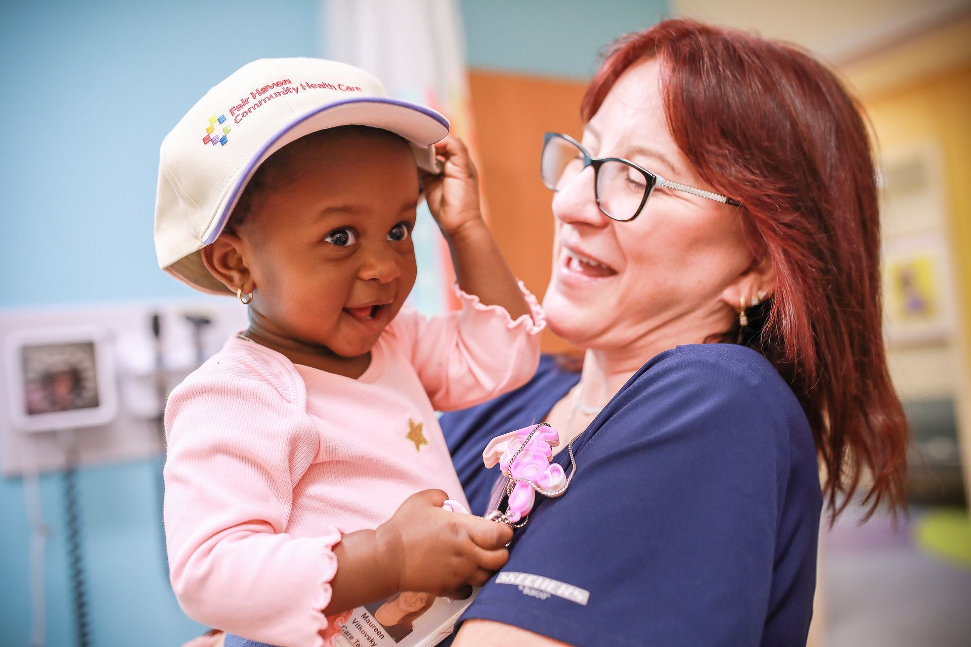 Woman holding a child wearing a white cap, both smiling in a bright indoor setting