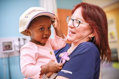 Woman holding a smiling toddler in a pink sweater and hat indoors