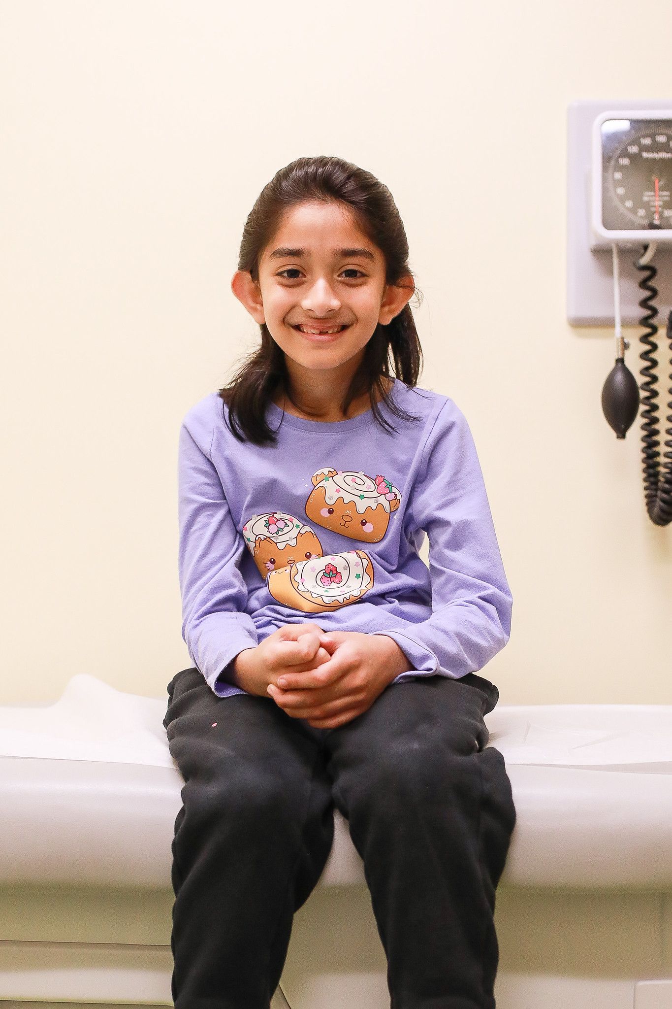 A smiling child in a long-sleeved purple shirt sits on a clinical examination table in a doctor's office.