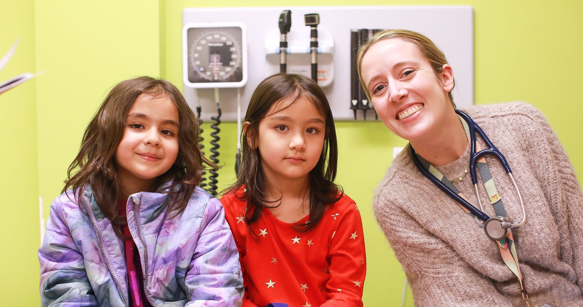 Three people smiling in a clinic exam room, including a doctor with a stethoscope and two children.