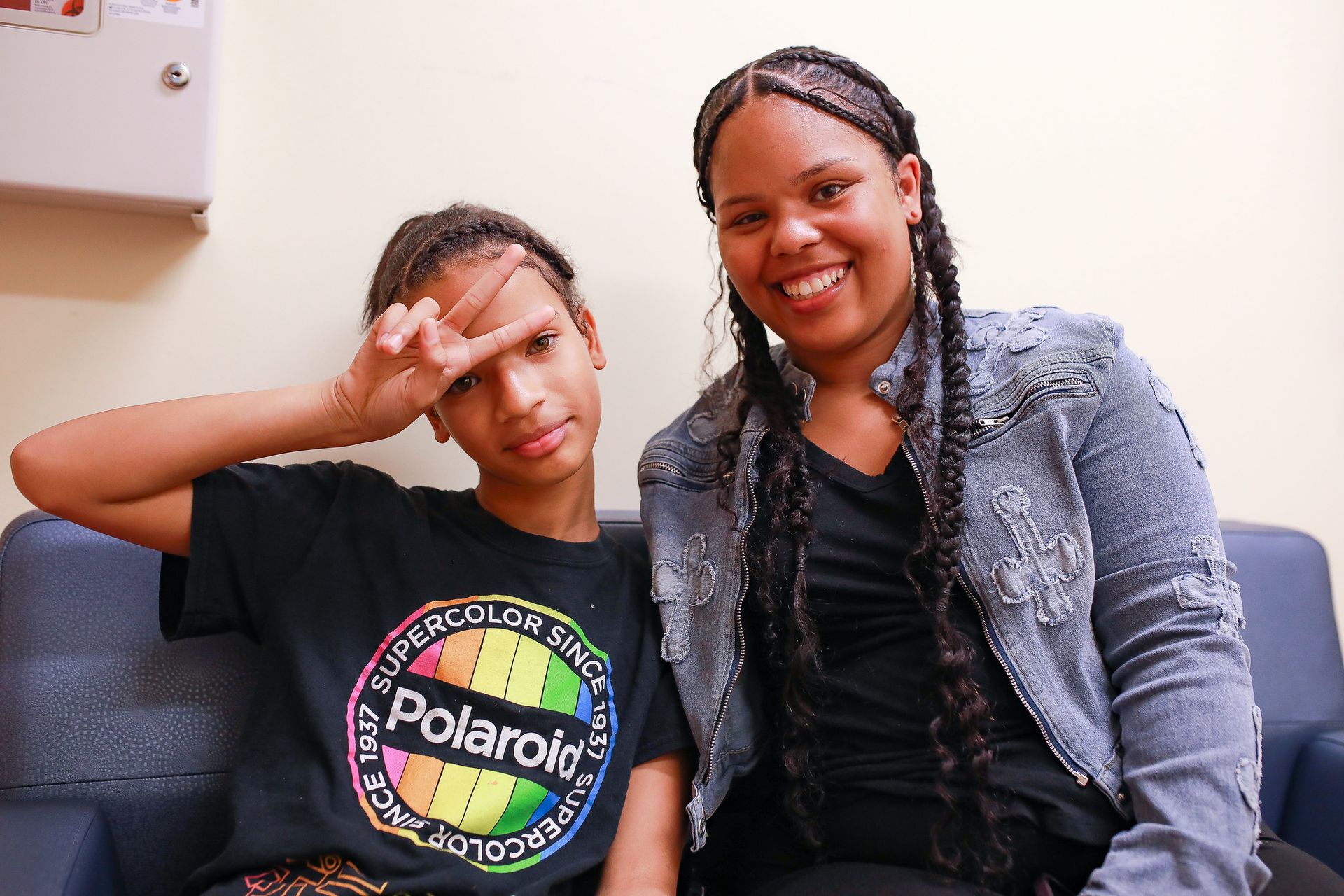 Two people sitting on a couch, one making a peace sign, smiling, and wearing a black Polaroid t-shirt and denim jacket.
