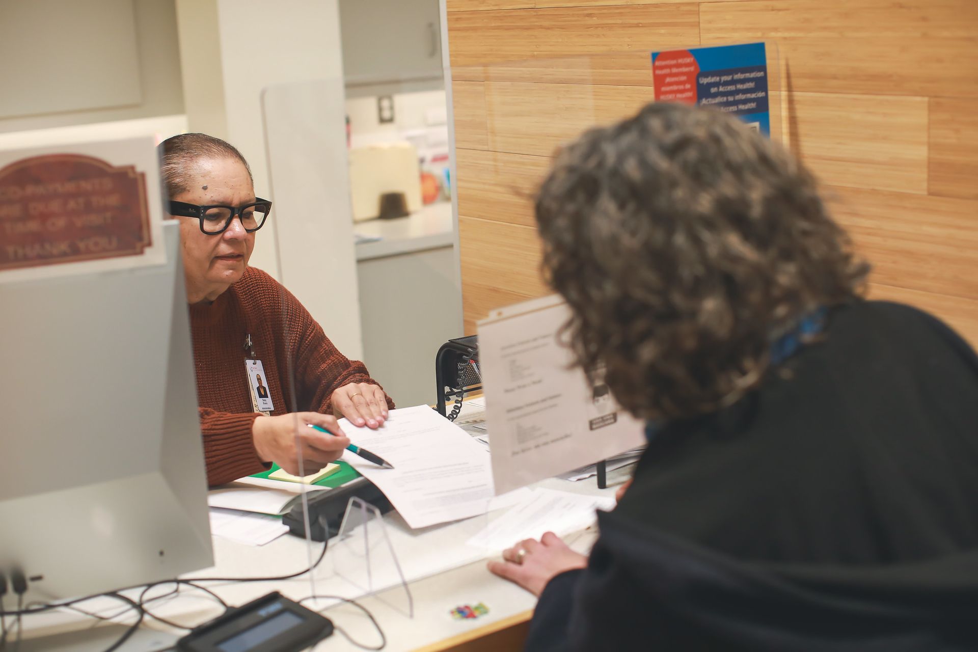 A person points to a document on a counter while speaking with another person at an office or clinic reception desk.
