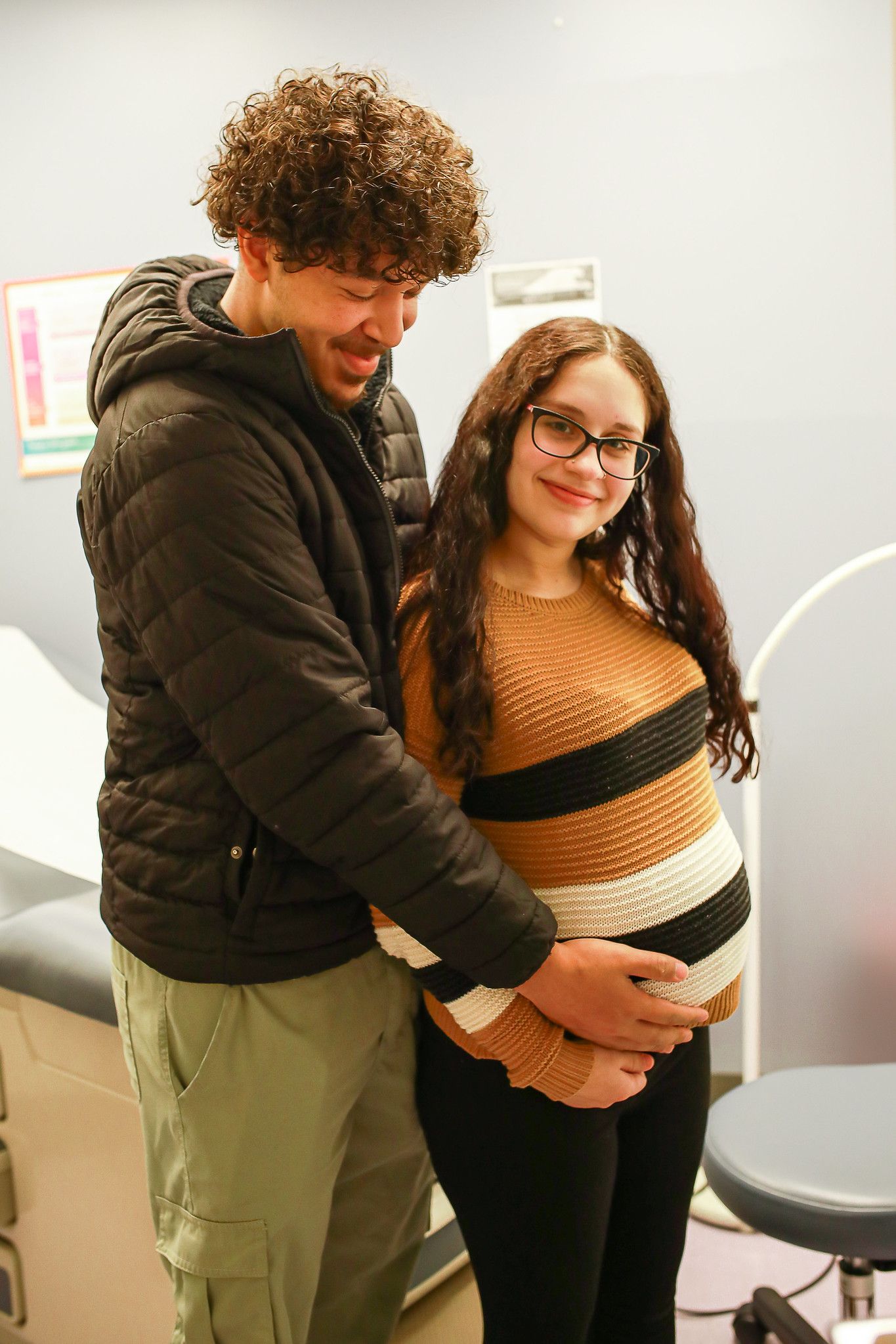 A smiling couple stands in a medical office, with the partner resting hands on the other person’s pregnant abdomen.