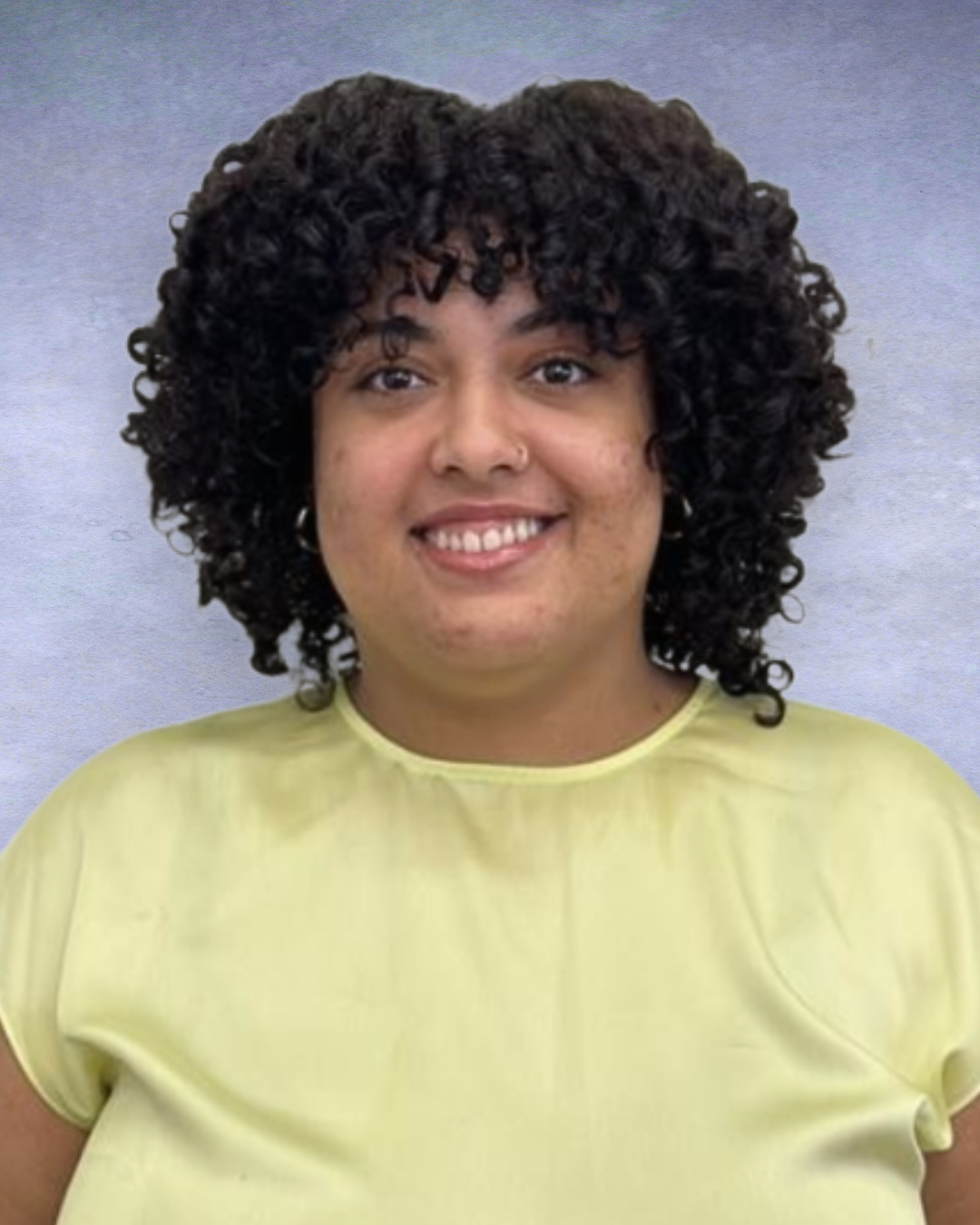 Smiling person with curly hair wearing a light yellow top against a gray background
