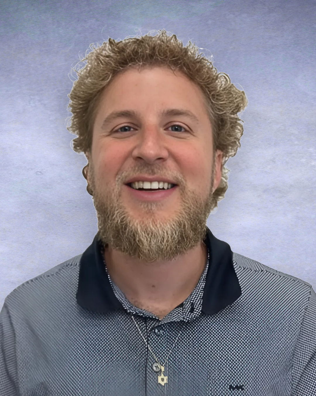 Smiling person with curly hair and beard wearing a blue collared shirt against a gray backdrop
