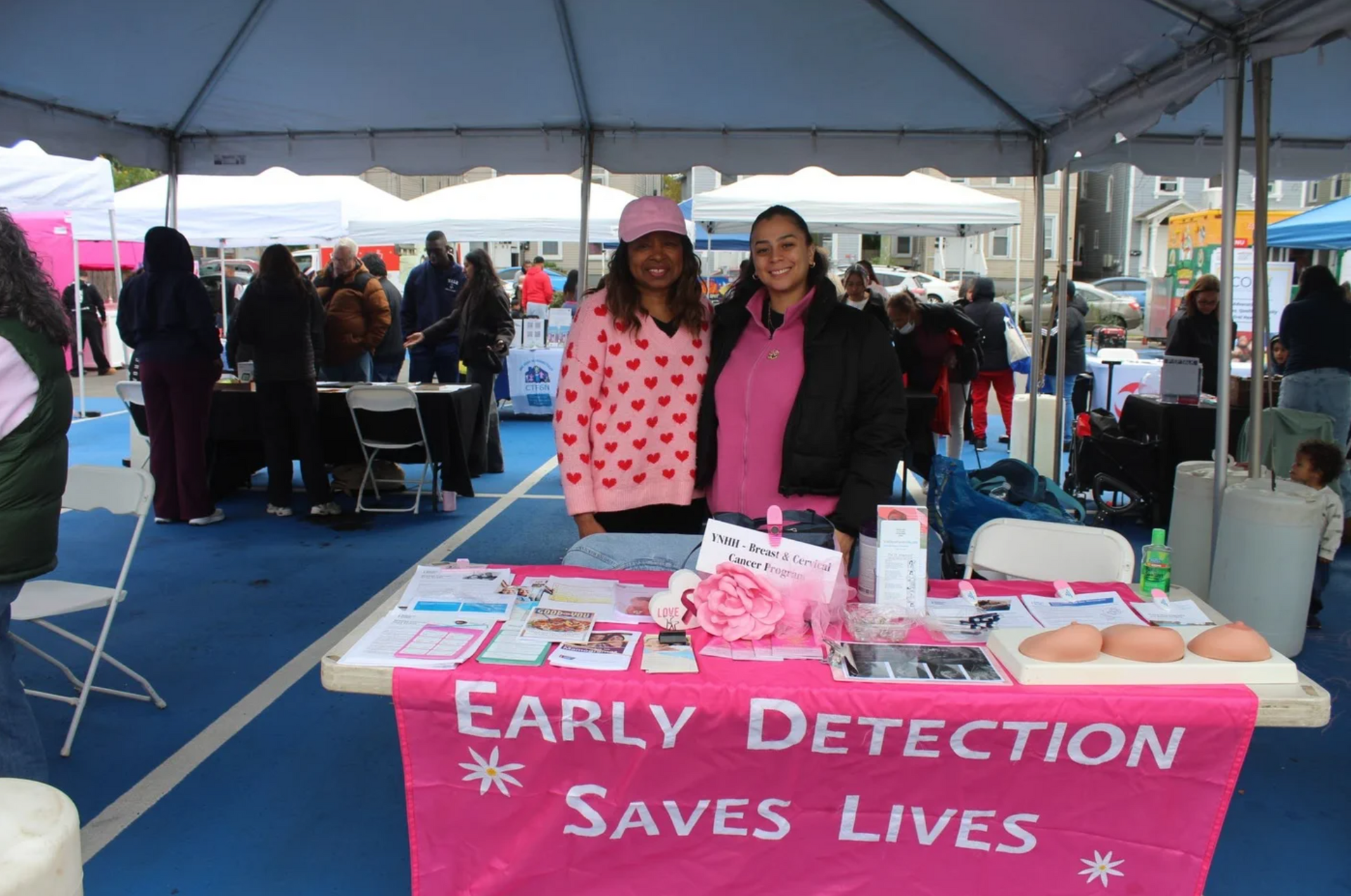 Two women at a pink breast cancer awareness booth under a tent, with sign reading “EARLY DETECTION SAVES LIVES”