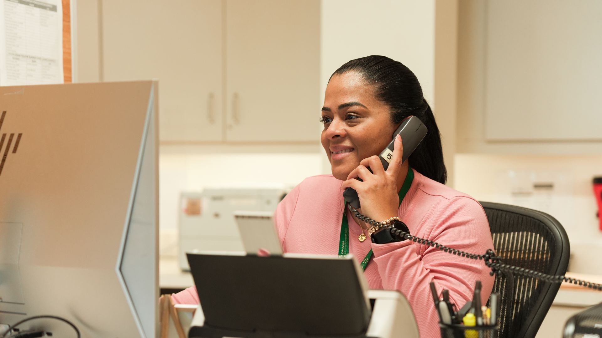 A person in a pink shirt speaks on a landline phone while sitting at a desk with a computer in an office.
