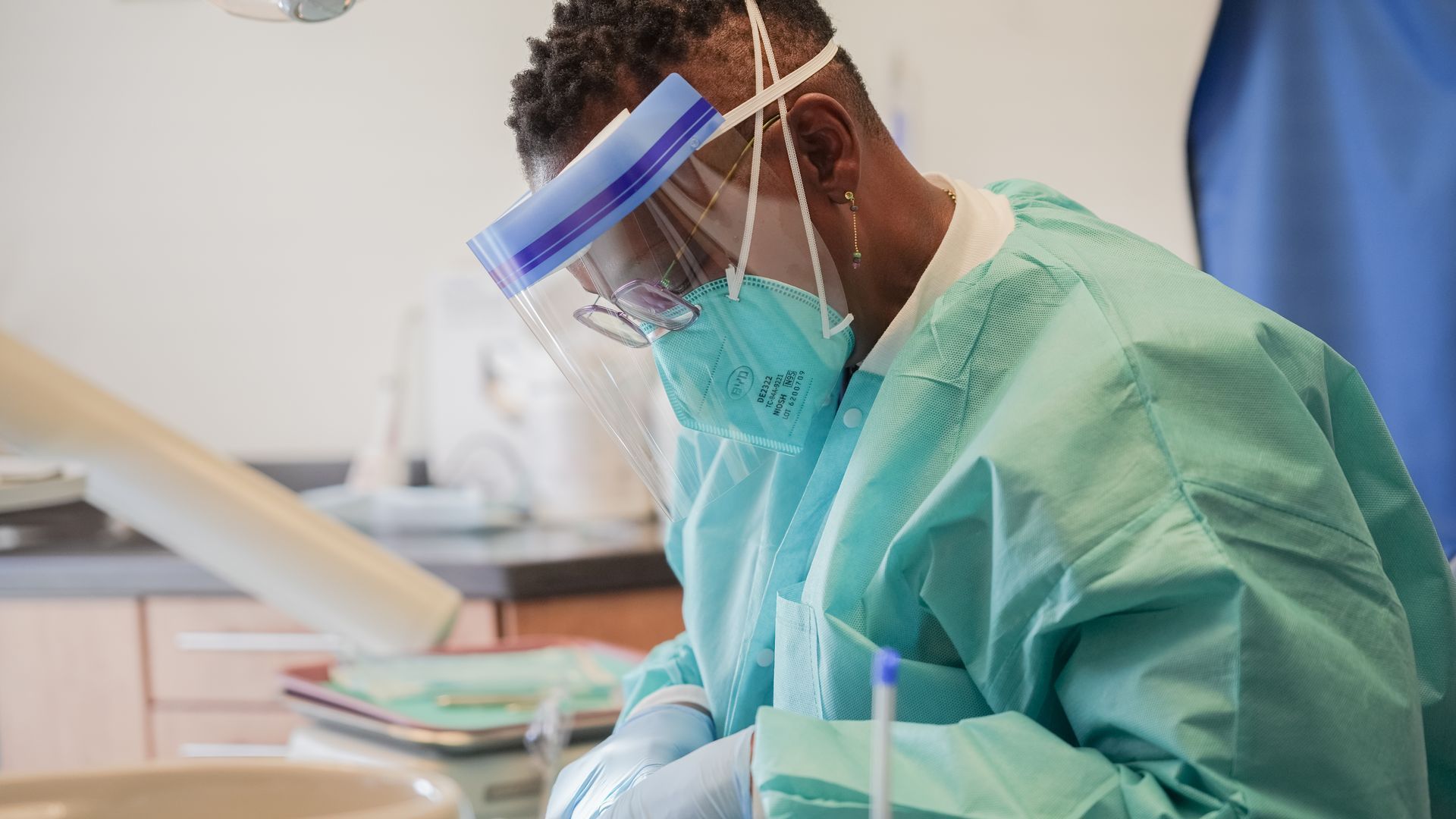A medical professional in a dental office wearing a face shield, mask, and gown, working over a patient.