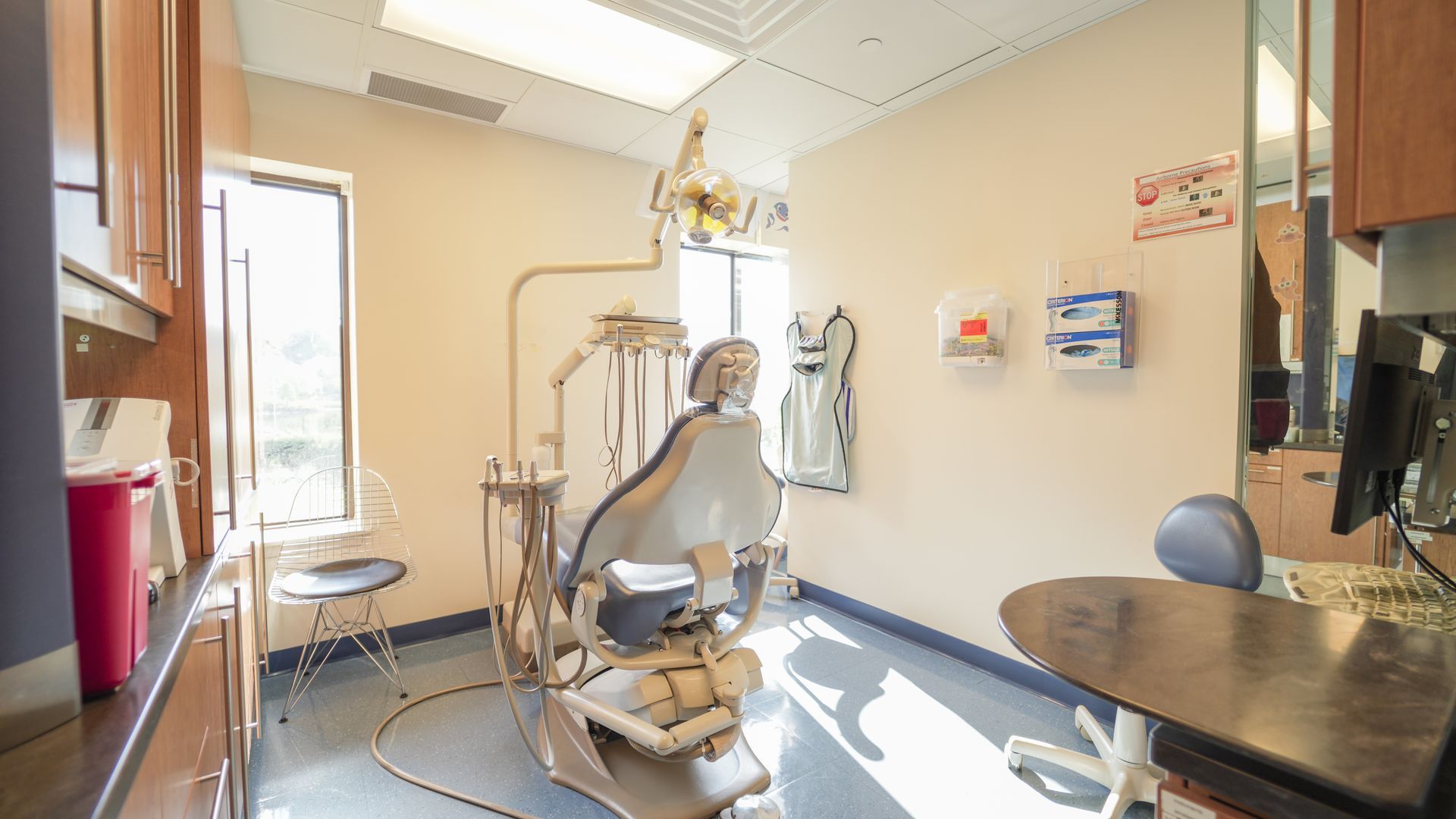 A bright, clean dental examination room featuring a centered patient chair, dental equipment, and a small workspace.