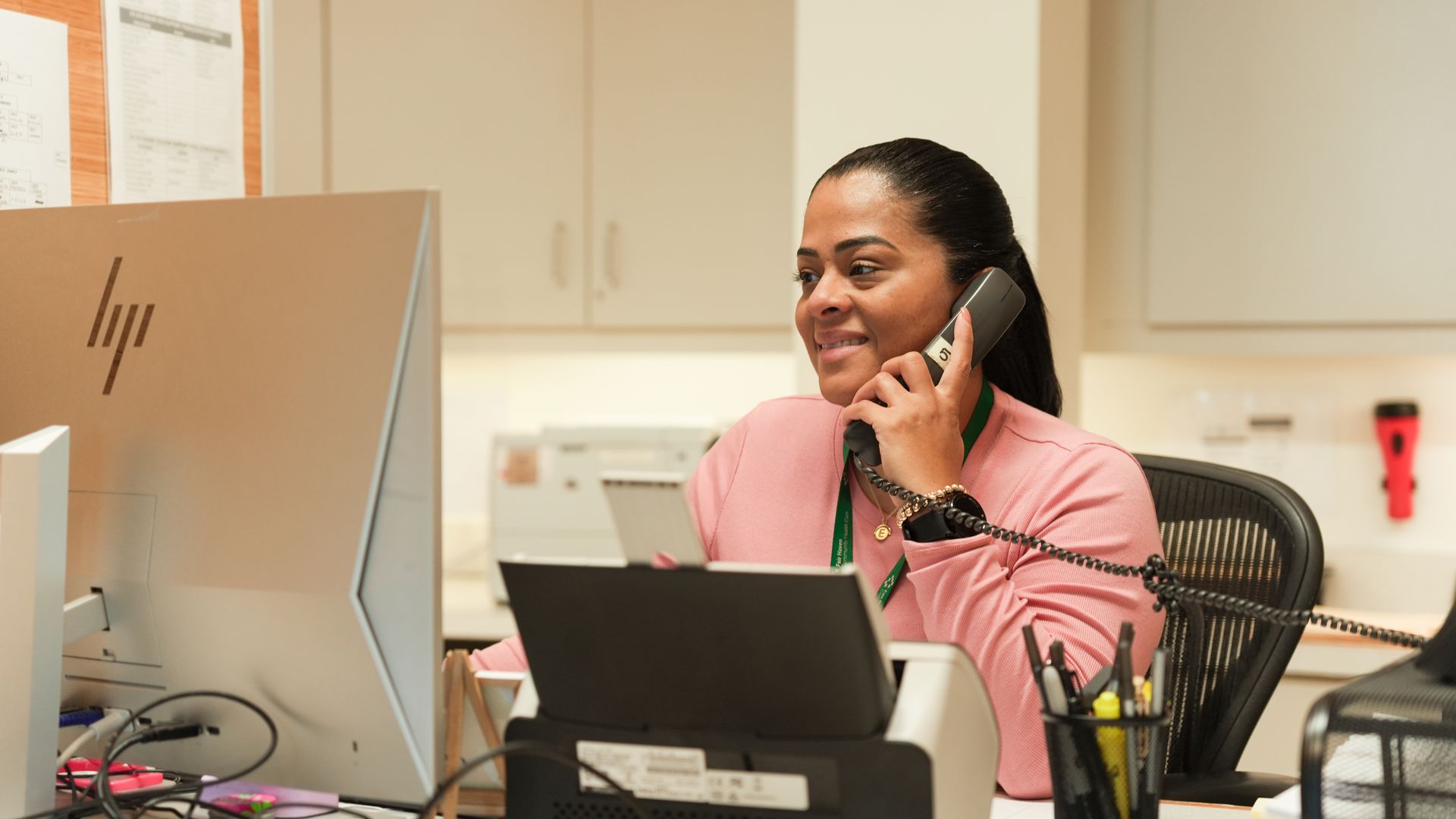 A person in a pink shirt sits at a desk using a telephone and looking at an HP computer monitor in an office setting.