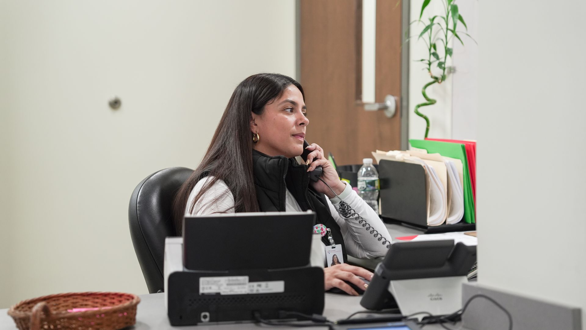 Woman seated at an office desk, smiling while on the phone beside a laptop and paperwork.