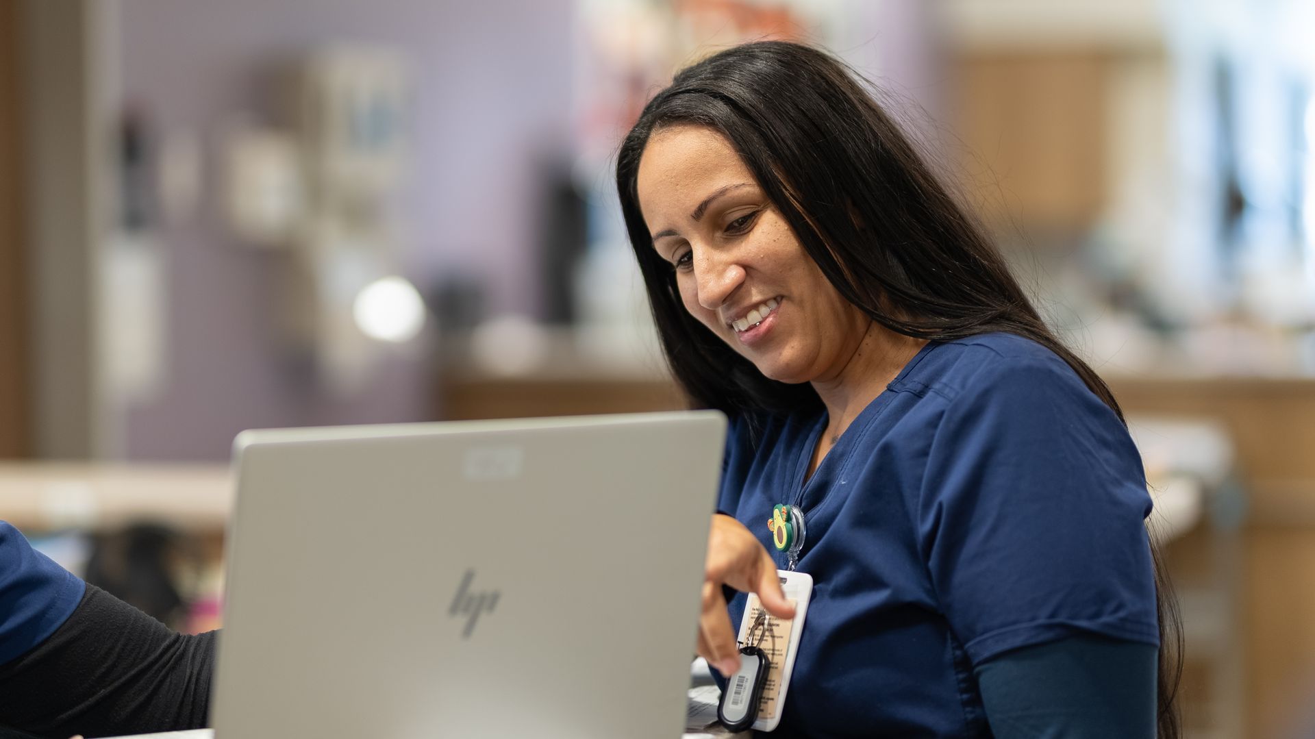 Woman in a navy shirt smiling while using a laptop in a bright indoor setting.