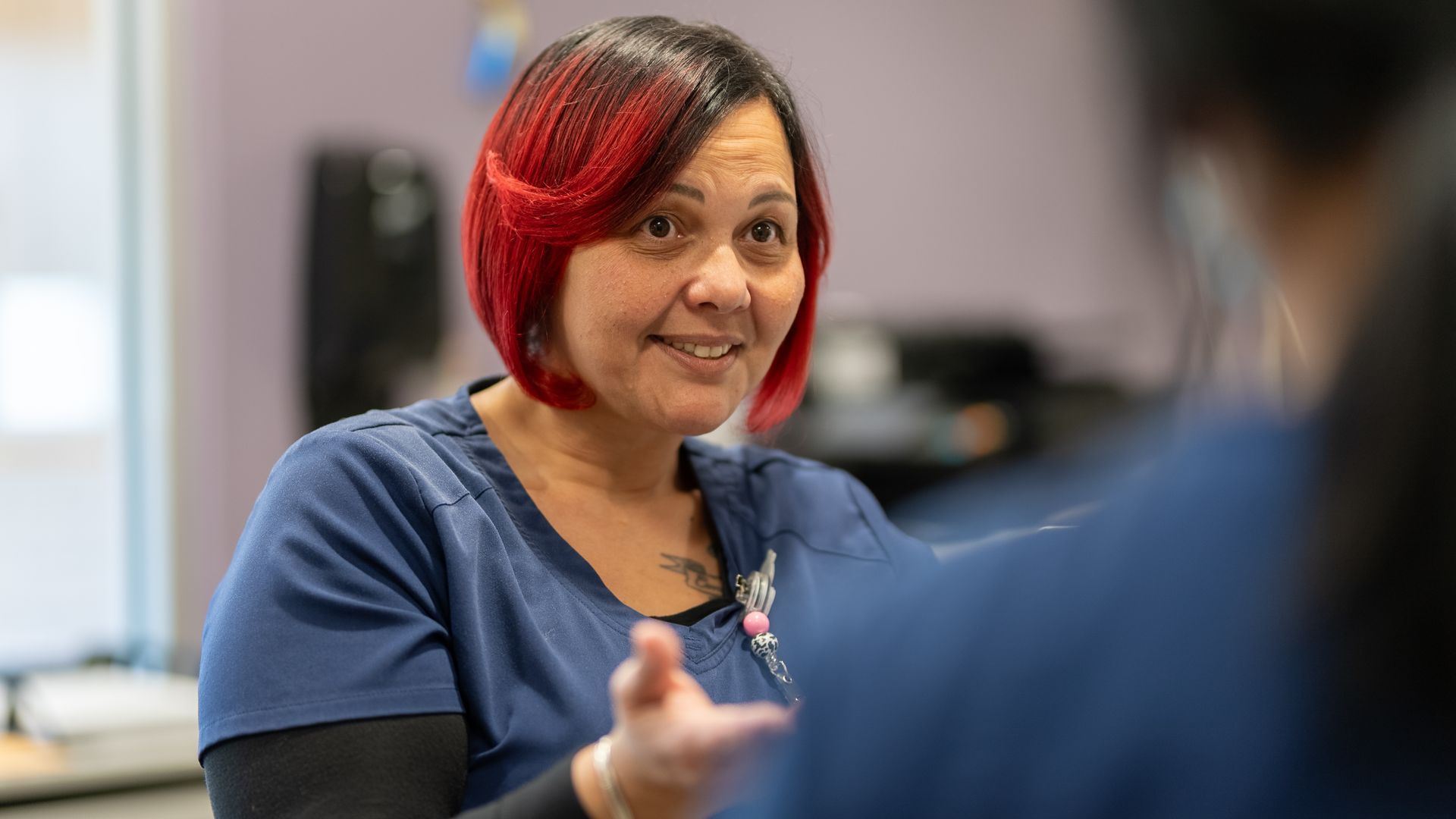 Woman with red hair speaking in an office, gesturing toward someone in the foreground.