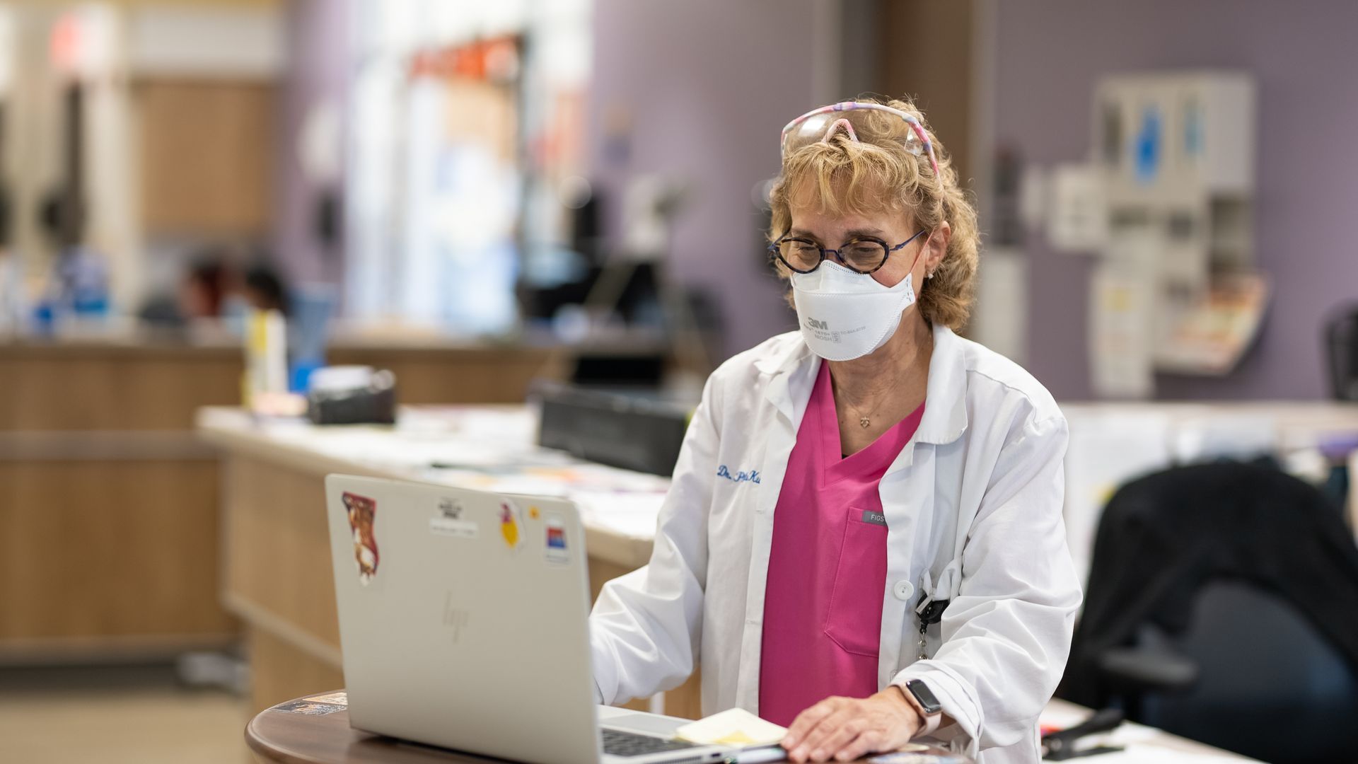 Masked healthcare worker in white coat and pink shirt working on a laptop in a hospital lobby