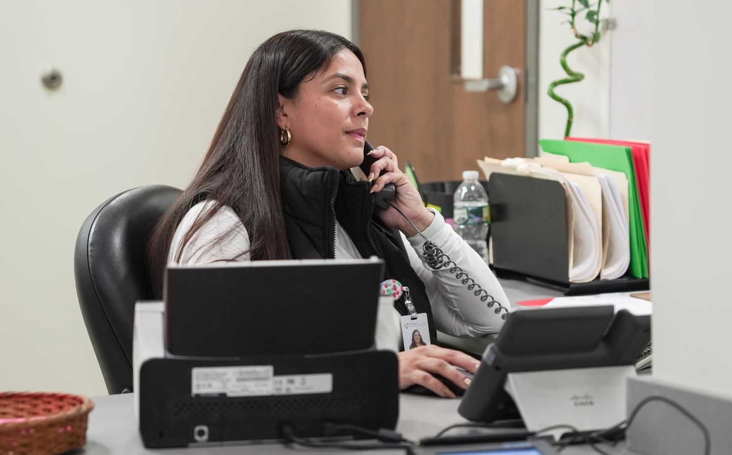 A person sitting at an office desk, talking on a landline phone while looking at a computer screen.