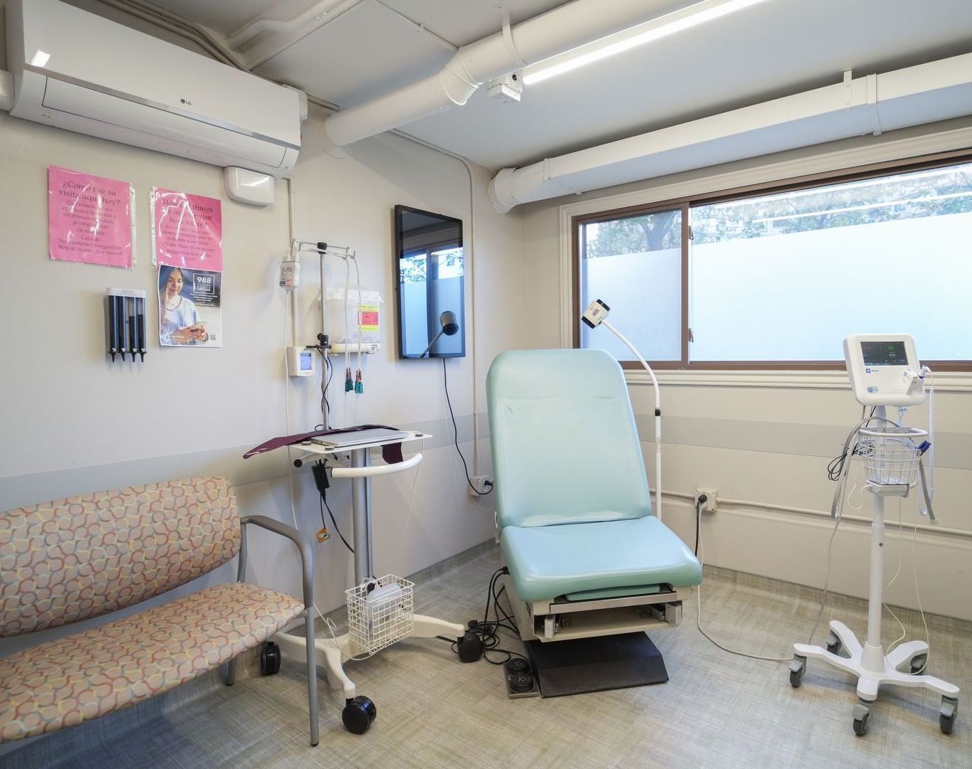 Medical exam room with a teal examination chair, bench seating, and monitoring equipment by the window.