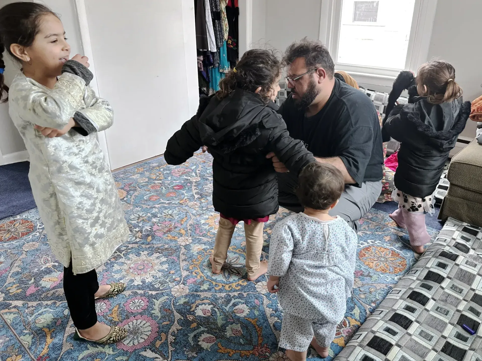 A man kneels on a floral rug to help a child into a black coat, surrounded by three other children in a room.