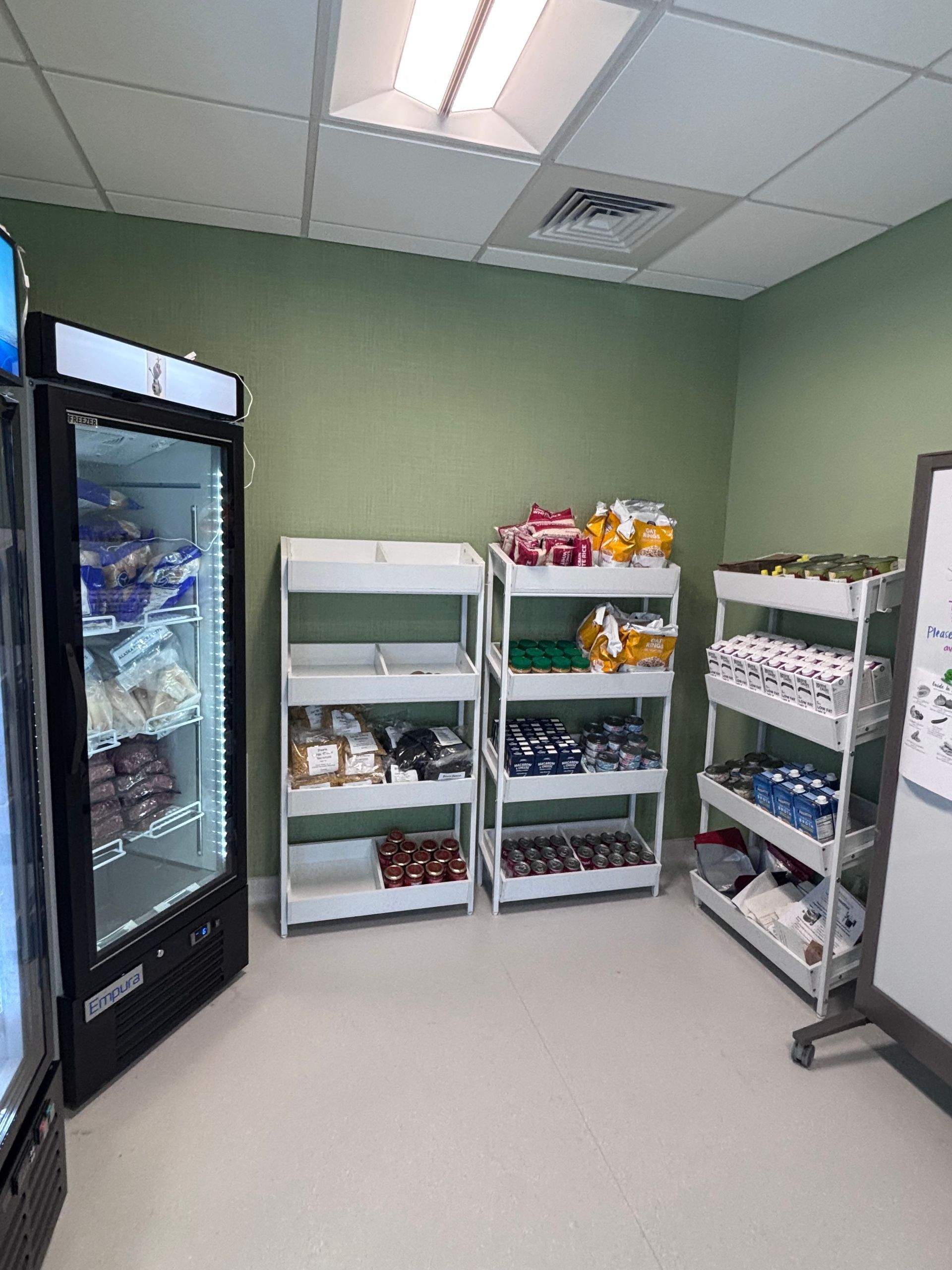Small storage room with shelves of packaged snacks, a glass beverage cooler, and a metal cart against green walls.