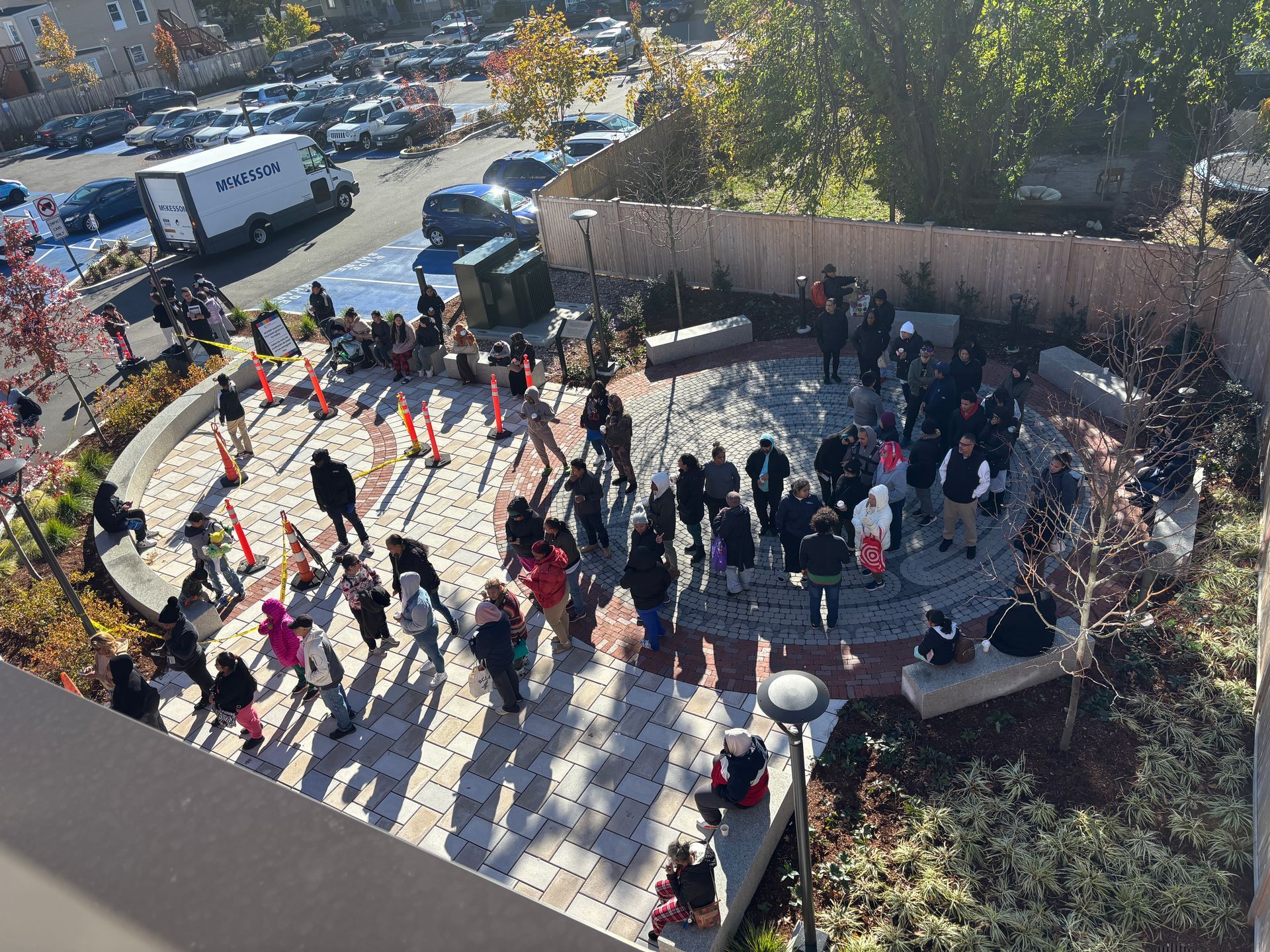 People gathered in a sunny courtyard, with long shadows and trees around a circular seating area.