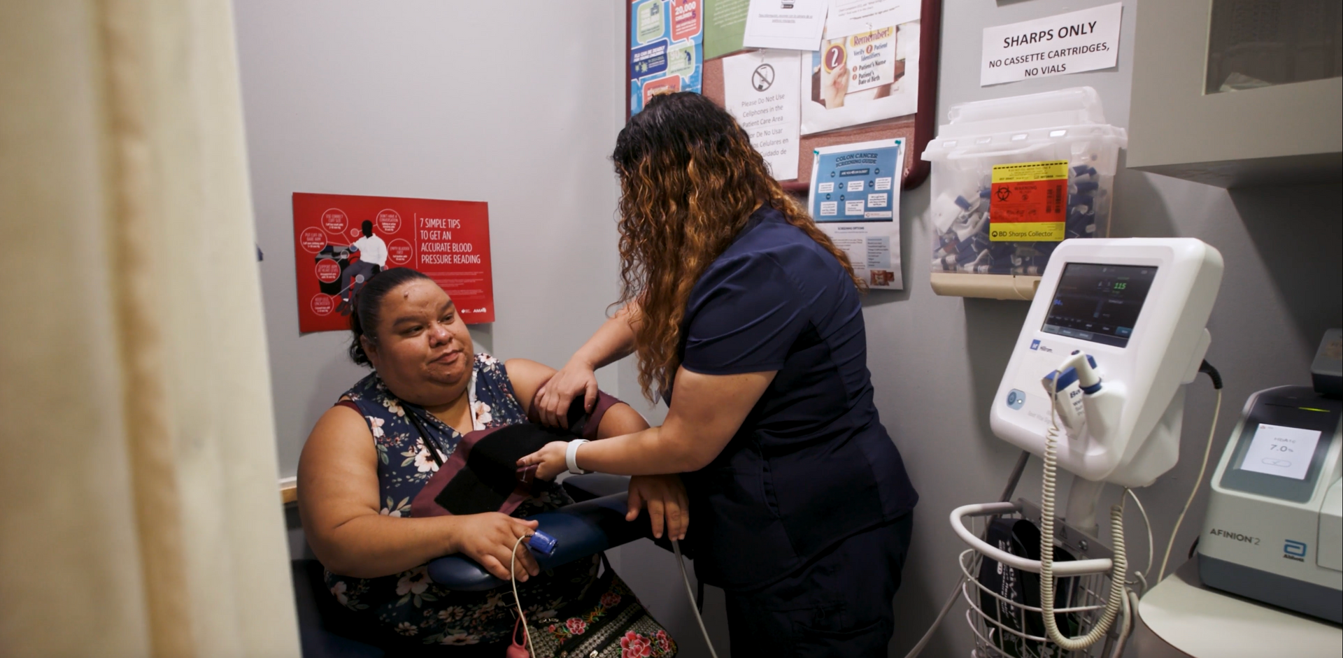 Clinician examines a seated patient in a small medical exam room, with equipment nearby.