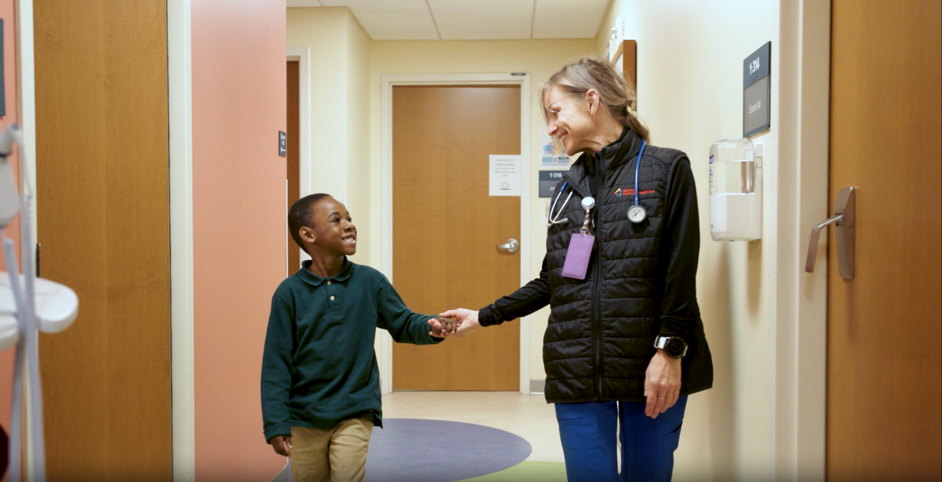Adult and child walking hand in hand in a hallway, smiling in a bright indoor setting.