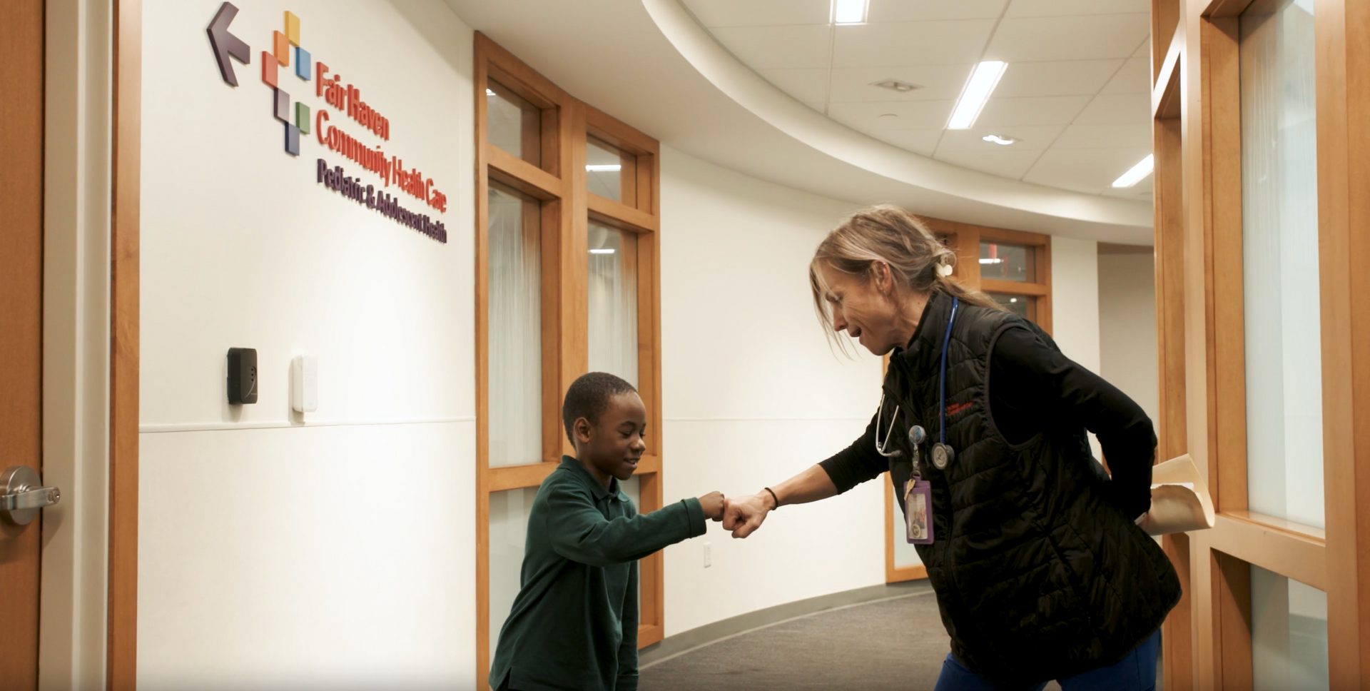 A woman greets a child with a handshake in a bright office hallway.