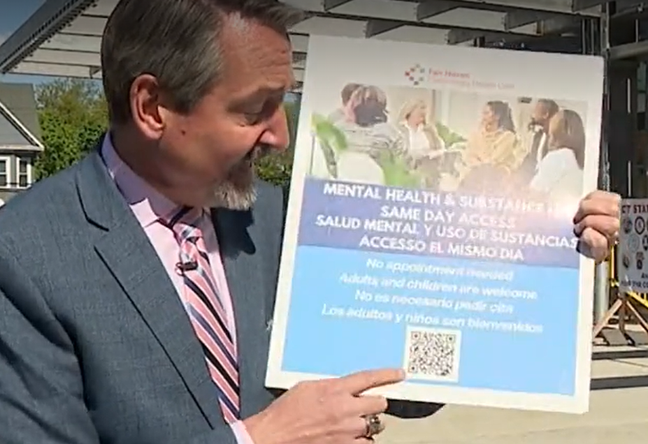 Man in suit points to a blue mental health awareness poster with multilingual text and a QR code outdoors