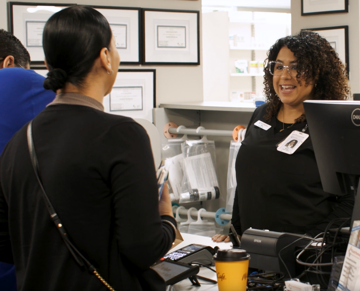 A staff member smiles while assisting a customer at a pharmacy service counter.