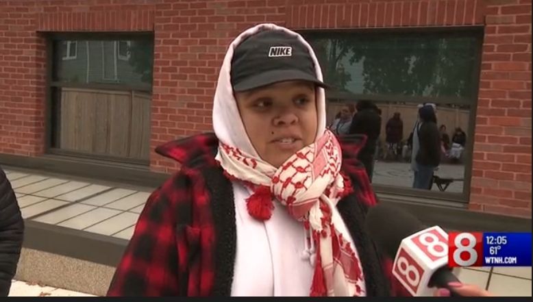 Person in red plaid coat and scarf speaks to news microphone outside a brick building