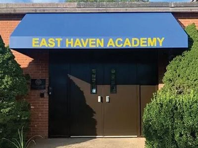 East Haven Academy entrance with blue awning over double doors and a walkway surrounded by greenery