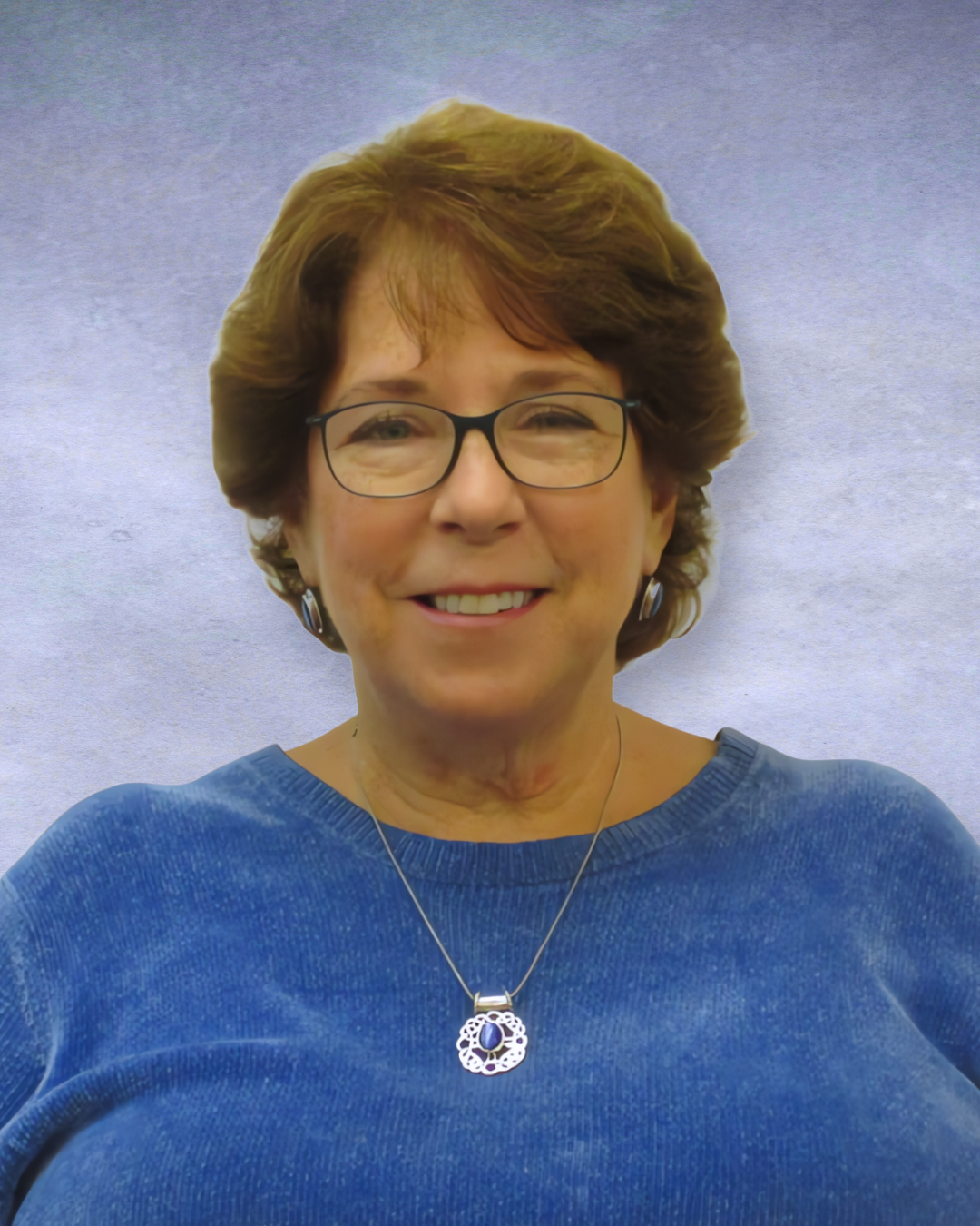 Portrait of a smiling person wearing glasses, a blue top, and a pendant necklace against a light purple background