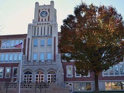 Historic brick school building with a clock tower and an American flag beside a large autumn tree