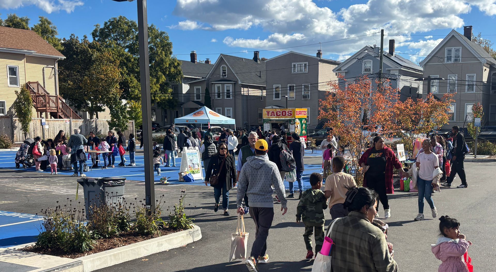 Crowd gathered at an outdoor street event near tents, blue crosswalks, and autumn trees under a sunny sky