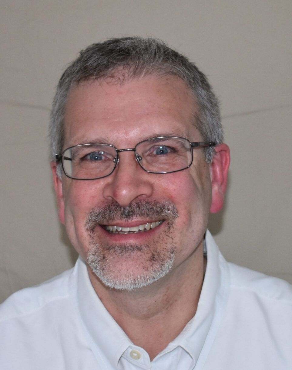 Smiling man with glasses and gray beard wearing a white collared shirt against a plain background