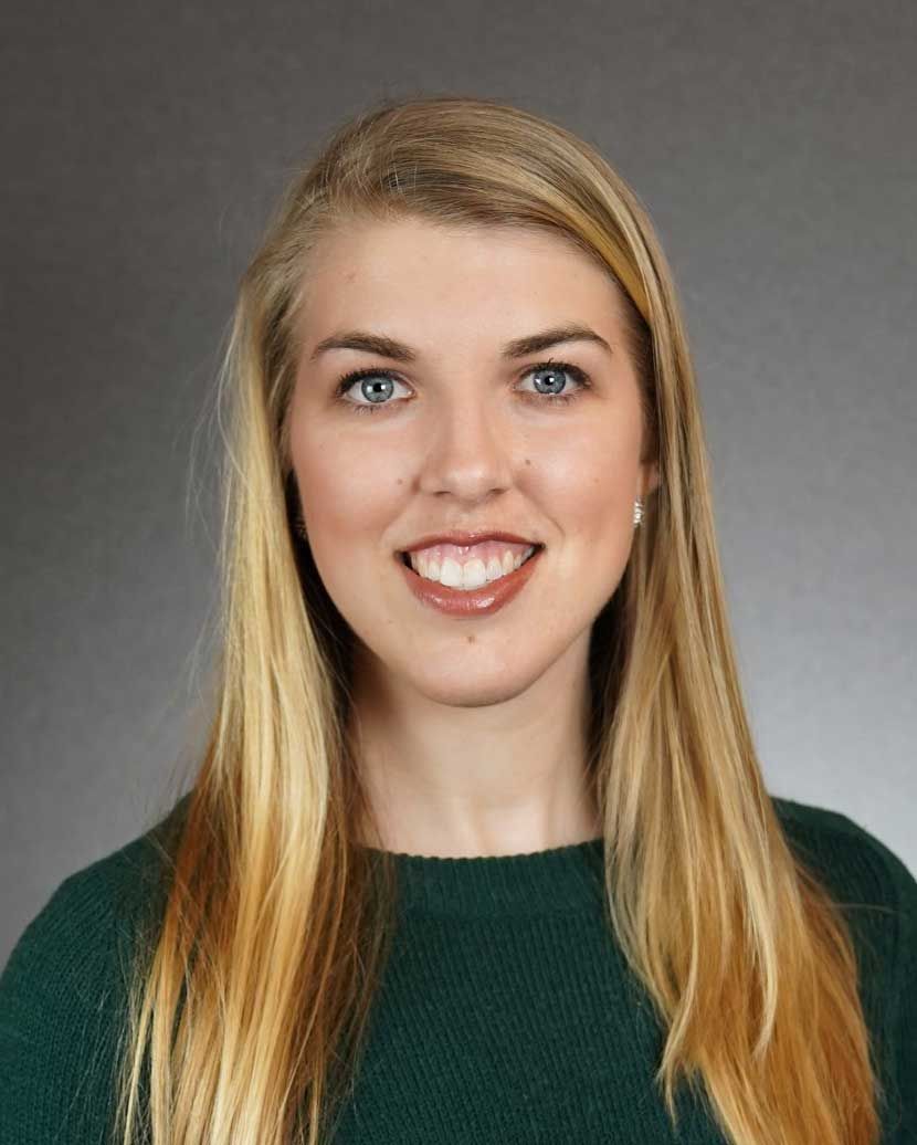Portrait of a smiling person with long blonde hair wearing a dark green sweater on a gray background