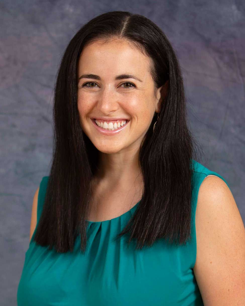 Smiling woman with long dark hair in a teal sleeveless top against a gray studio backdrop