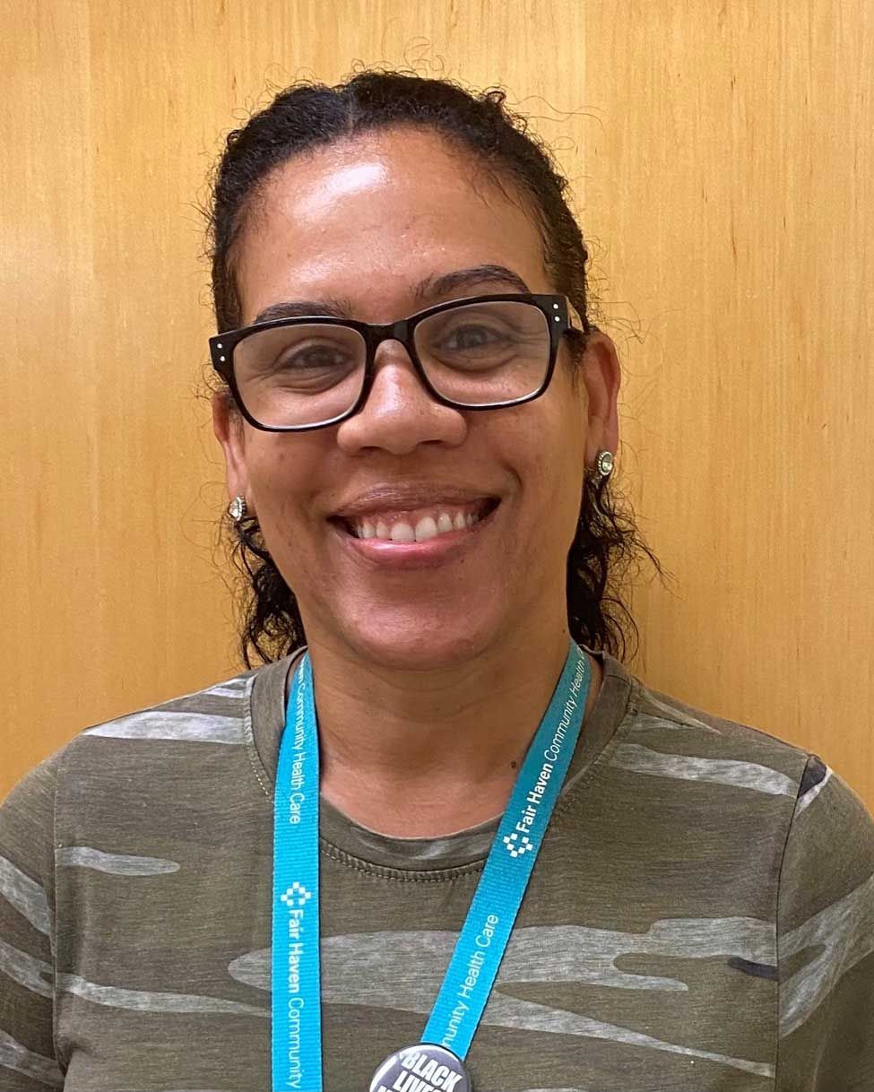 Smiling person wearing glasses and a blue lanyard against a wooden wall.