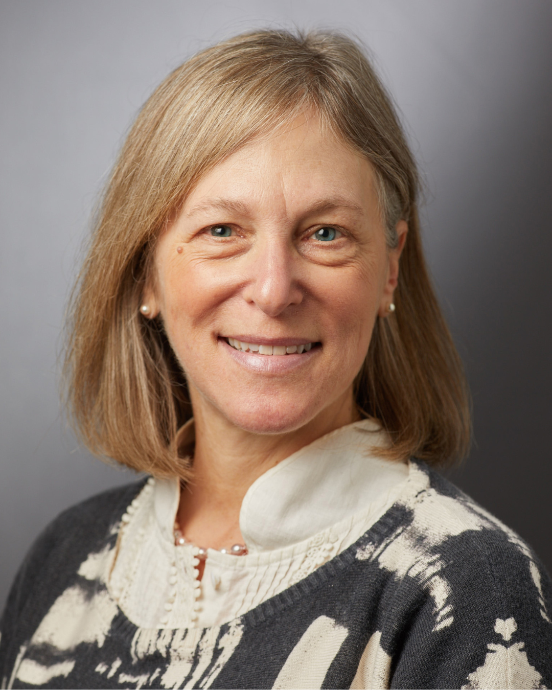 Smiling person in a cream blouse and dark patterned sweater against a gray background