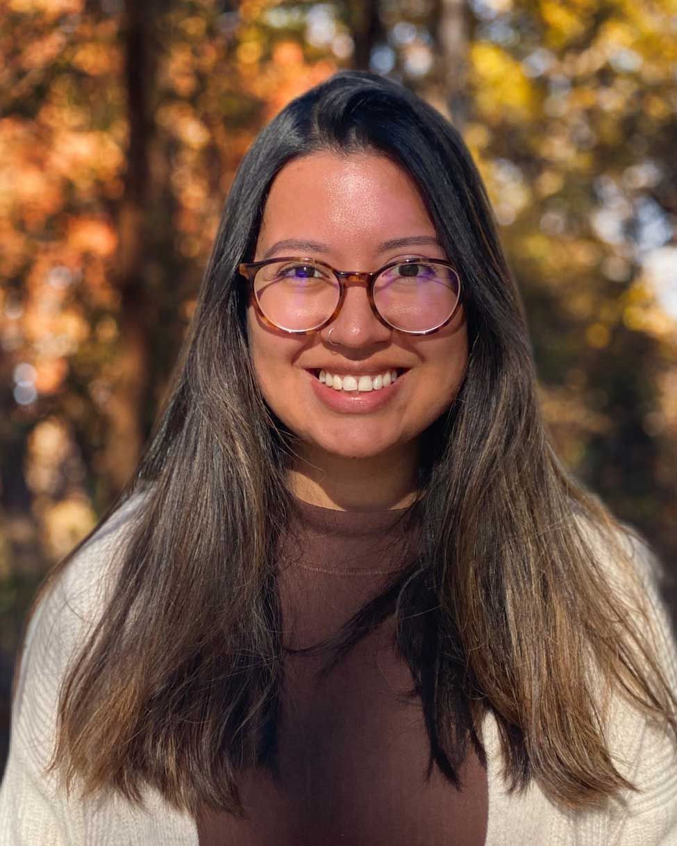 Smiling person with glasses and long hair outdoors in autumn, wearing a brown sweater.