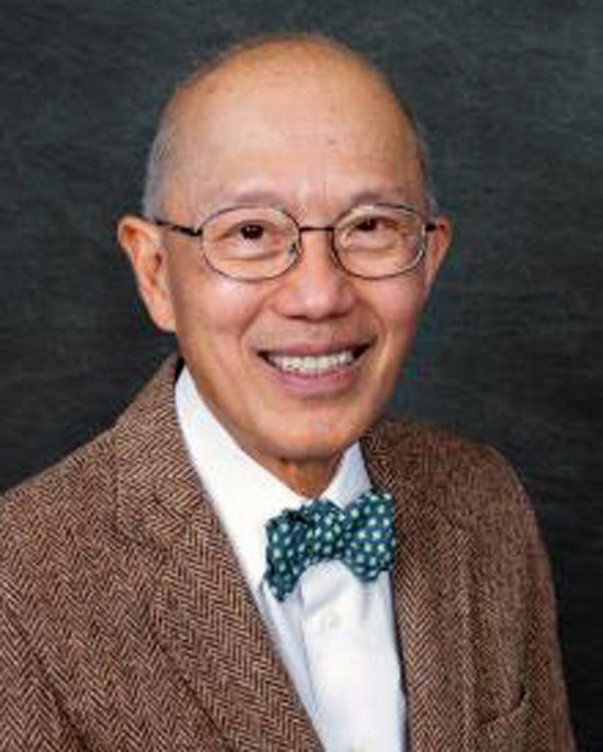 Portrait of a smiling man in glasses, wearing a brown sweater and green bow tie against a dark background