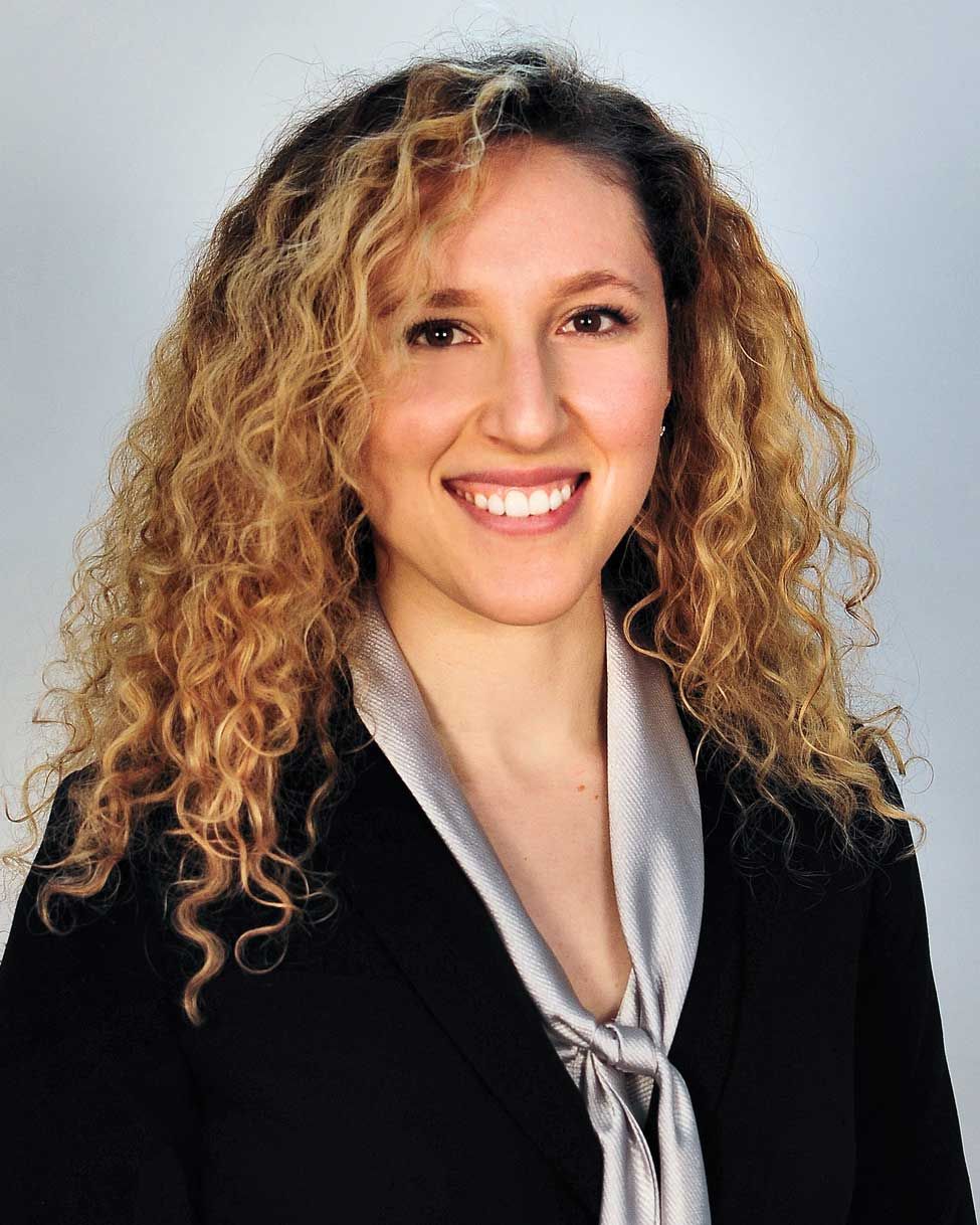 Smiling person with curly blonde hair wearing a black blazer and silver blouse against a gray background