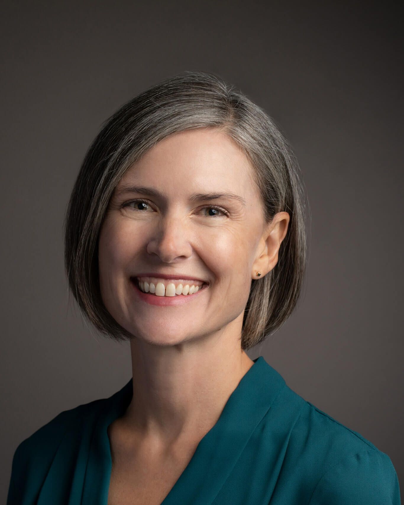 Smiling person with short gray hair wearing a teal blazer against a dark background