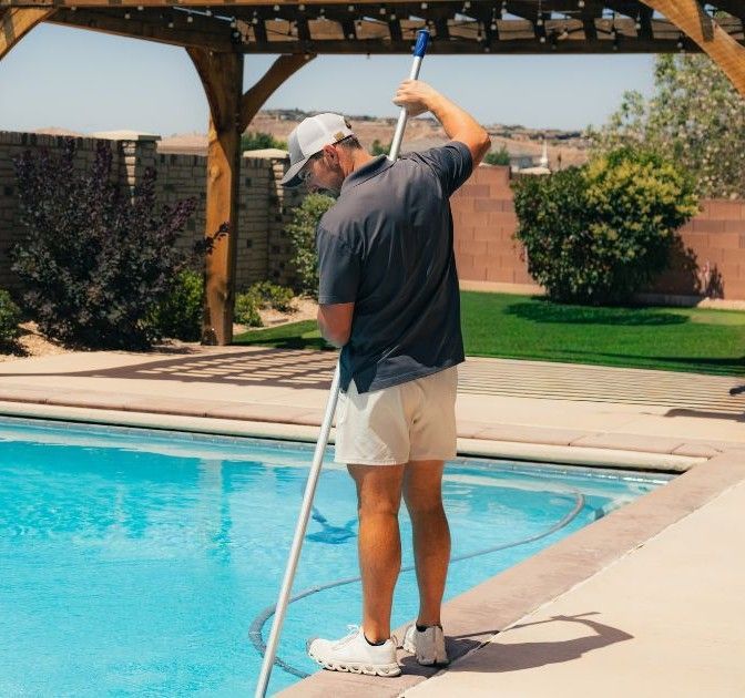 Person cleaning a pool with a long pole beside a backyard swimming pool