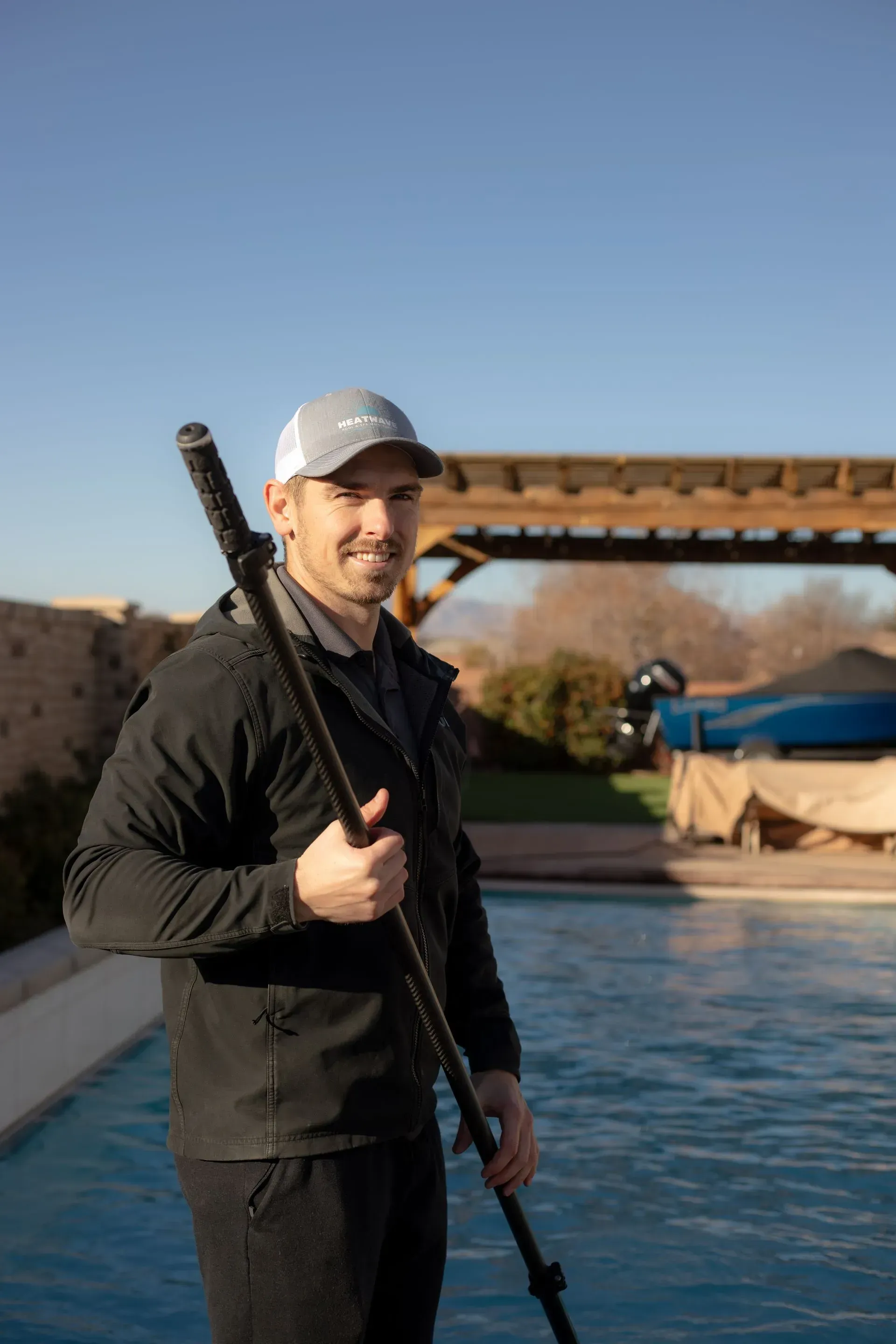 Person holding a long pole beside a pool at sunset, with a bridge in the background