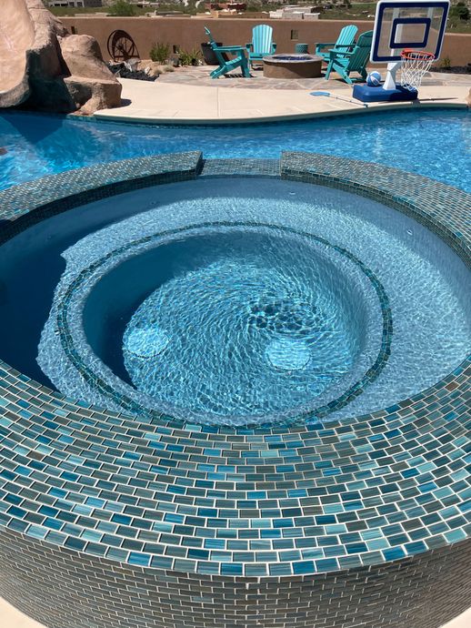 Blue mosaic hot tub with bubbling water beside a swimming pool and patio chairs.